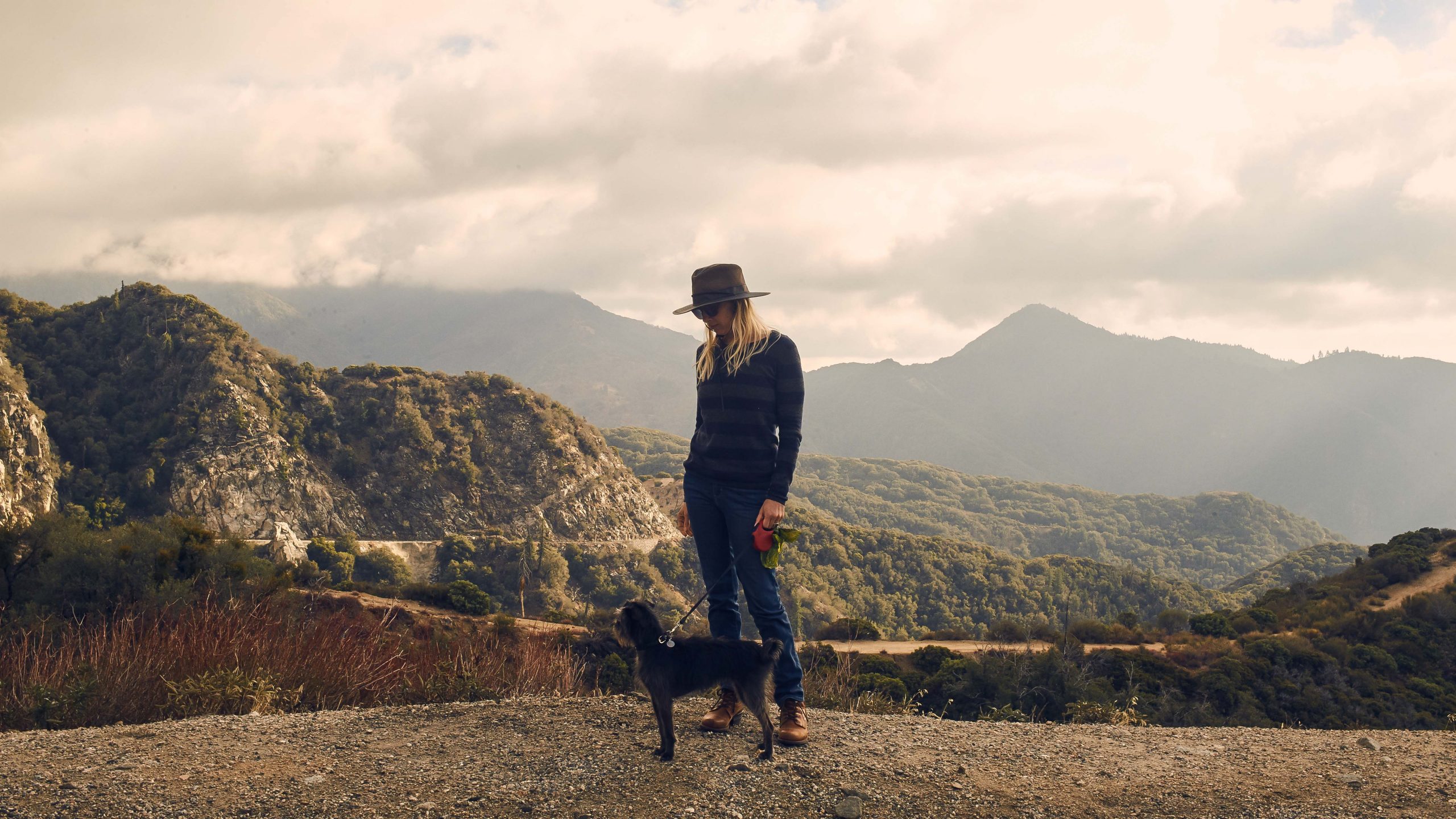 On a gravel path with mountainous terrain and cloudy skies in the background, someone stands wearing a hat, sunglasses, and a striped sweater. They hold a leash attached to a small black dog. The setting hints at a vacation rental in Big Bear Lake.