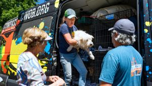 A woman lifts a small white dog from a van marked "Compassion Without Borders." Nearby, two people—one in a plaid shirt and the other in a blue staff shirt—watch. The cages inside the van and trees in the background suggest that this organization rescues dogs from Mexico.