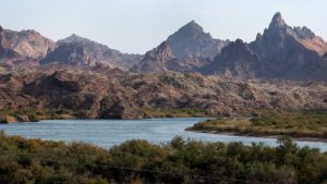 Moabi Regional Park offers a diverse landscape for you and your dog to explore. The centerpiece is a winding river edged by lush green vegetation, creating a striking contrast against the rocky, arid surroundings. Jagged mountains rise in the background, set against a clear sky. The terrain beyond the riverbanks is dry and barren, making the riverside trails an inviting option for walks with your pet.