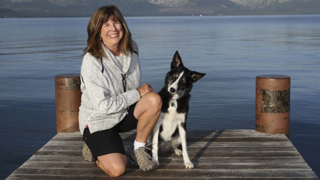 A woman with shoulder-length hair, dressed in a gray sweater and black shorts, kneels on a wooden dock by a lake. She smiles with purpose, her arm around a black and white dog sitting next to her. Behind them, the calm water and distant mountains set the scene for their time together.