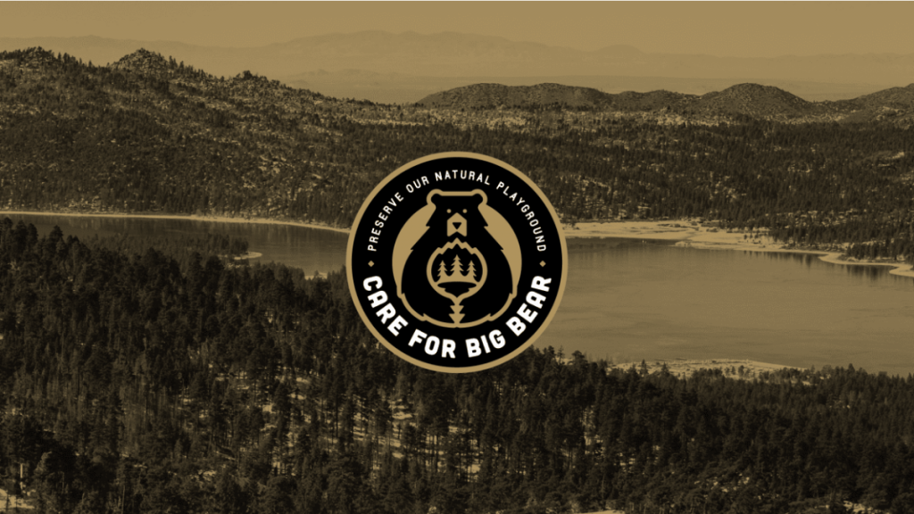 An aerial image captures a forested mountain range encircling a large lake, tinted in sepia tones. In the middle of the photo, a circular logo with an illustration of a bear and pine tree contains the words "Preserve Our Natural Playground - Stay and Care For Big Bear.