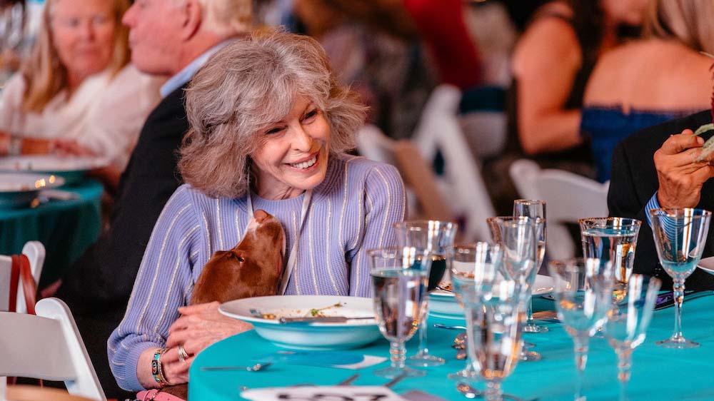 A woman with gray hair sits at a table set with glassware and plates during the Tails by Twilight event. She smiles, wearing a purple striped top, while gently holding a brown dog that looks up at her. In the background, people are eating and enjoying their meal.