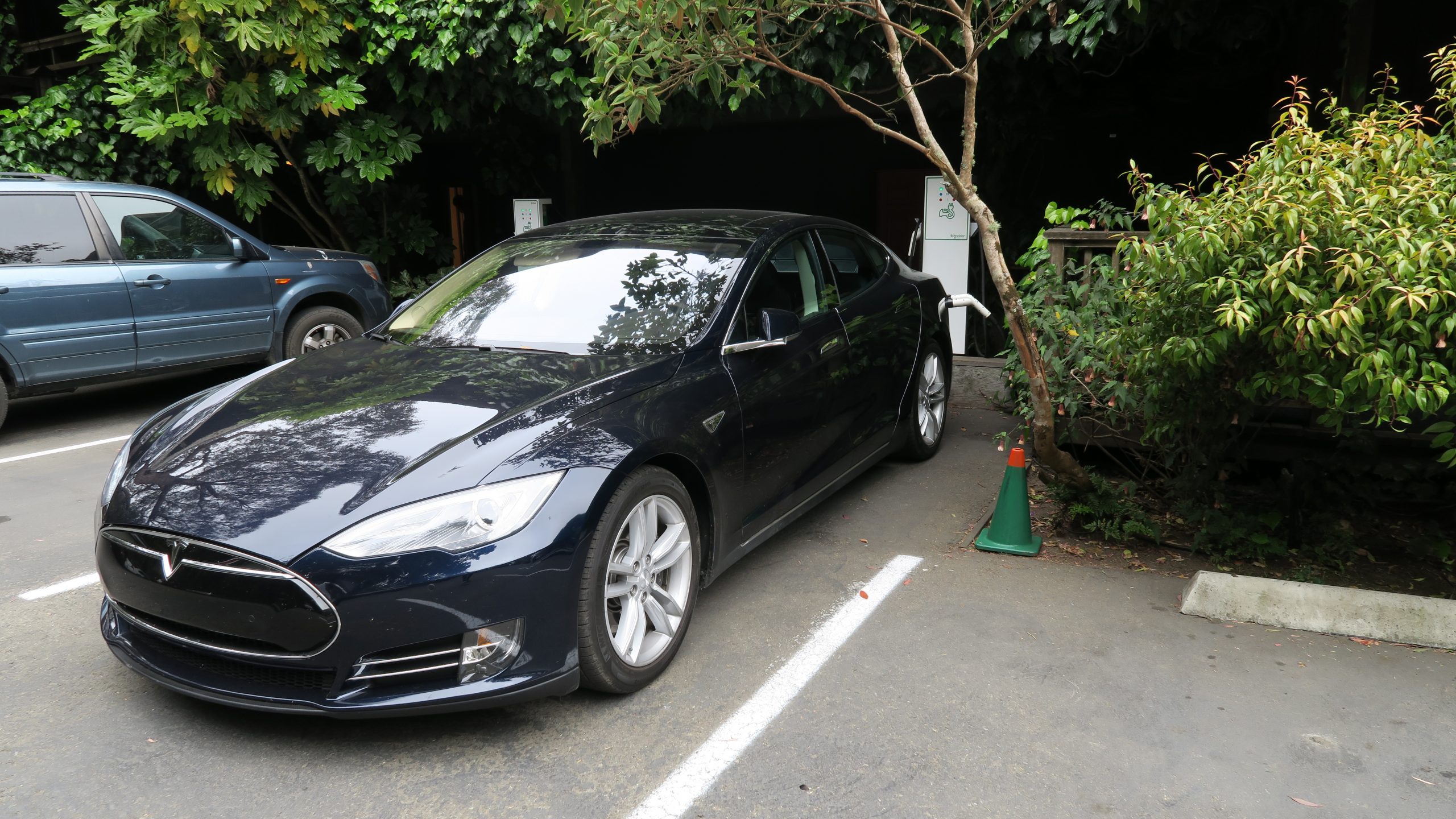 A black Tesla is charging at an electric vehicle station outside the Stanford Inn. The car’s charging cable is connected, and green plants surround its parking spot. Another vehicle is parked to the left, and a small green traffic cone sits on the right side of the Tesla.