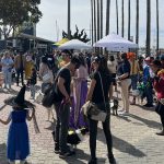 A lively outdoor, seasonal event where attendees in costumes like witches and butterflies gather. Numerous tents are set up, surrounded by trees and buildings. It's a dog-friendly occasion with some dogs dressed up and interacting cheerfully with the people.