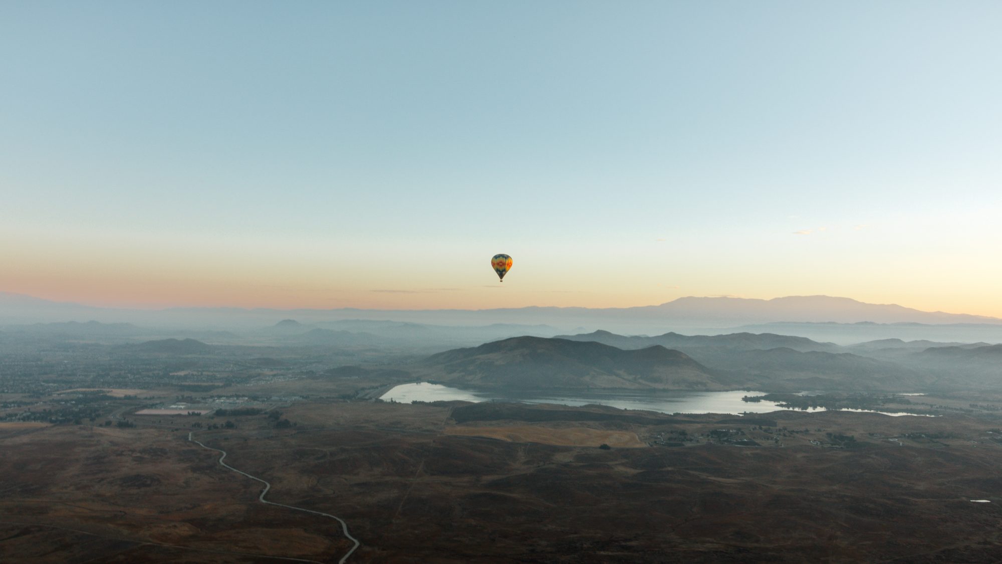 A hot air balloon drifts over a broad landscape with mountains, Lake Skinner, and wide plains during sunrise or sunset. The sky is clear, offering an unobstructed view of the peaceful scenery from above.