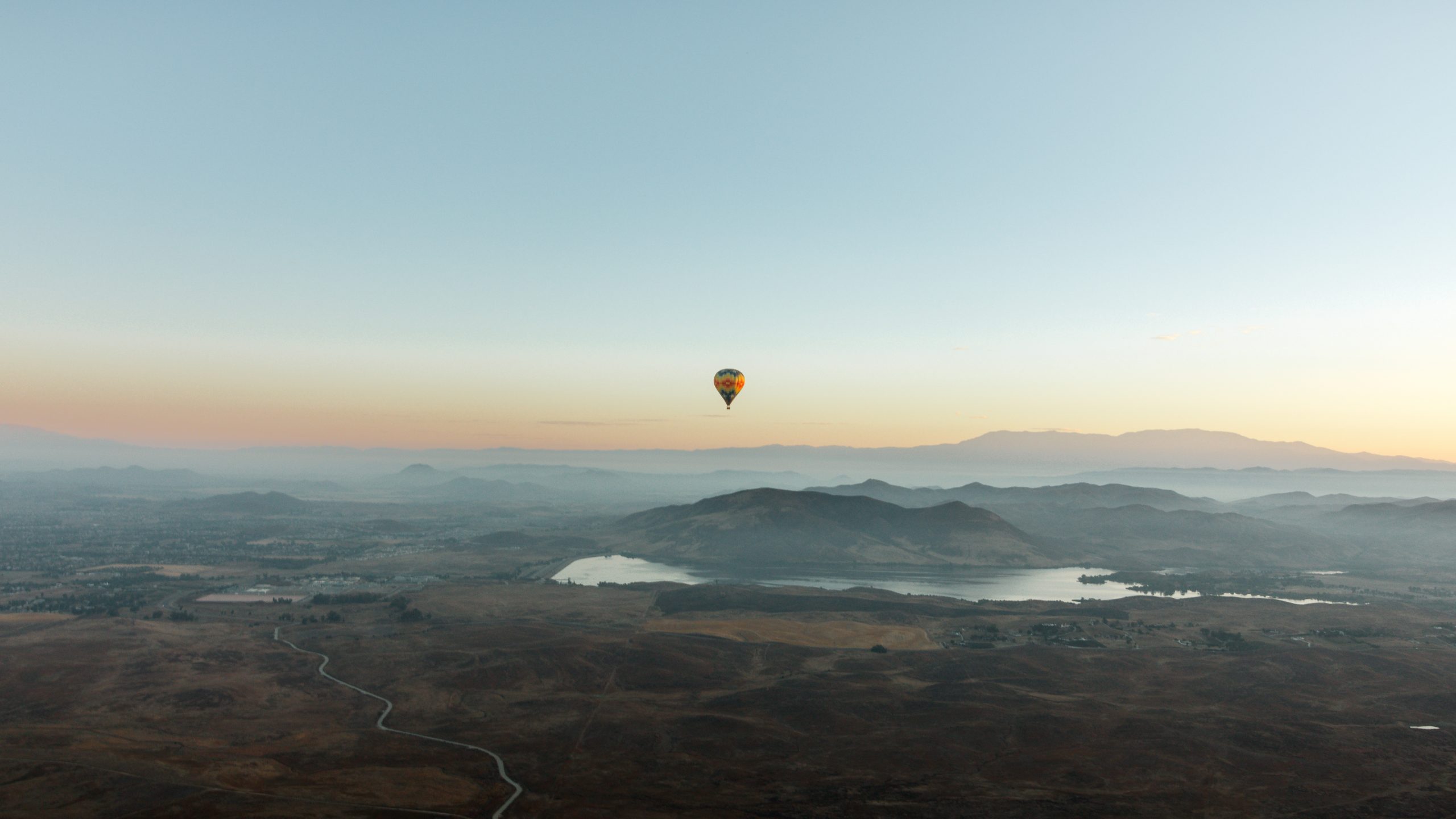 A hot air balloon drifts over a broad landscape with mountains, Lake Skinner, and wide plains during sunrise or sunset. The sky is clear, offering an unobstructed view of the peaceful scenery from above.