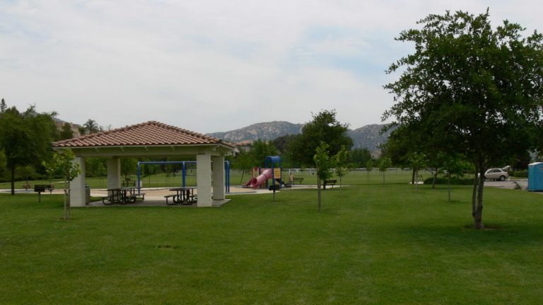 A vibrant dog park bathed in sunlight features abundant greenery with mature trees, offering ample shade for leisurely outdoor activities. In the distance is a well-equipped playground that presents an additional activity area. An inviting picnic table spreads out nearby, ideal for enjoying meals al fresco or taking relaxing breaks from play. A neatly-placed trash bin aids in maintaining the park's cleanliness for everyone's enjoyment. Dominating the scene is a generously wide dirt path gracefully curving through the expanse of grass, promising exploratory walks and lively fetch games.