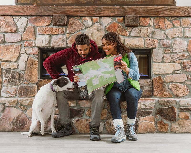 A man and woman sit on a stone bench in front of a fireplace, studying a map with smiles on their faces. The man pets a black-and-white dog that sits close by. Dressed in casual warm clothing, they look eager as they plan their fall visit to Big Bear for some hiking.