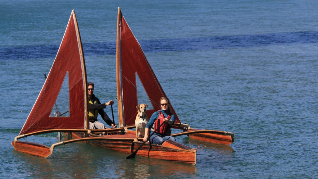 Two people and a dog are in a small wooden boat with red sails on calm water near the Stanford Inn. The person at the front holds a paddle, and the person at the back steers. The dog sits between them, gazing ahead.