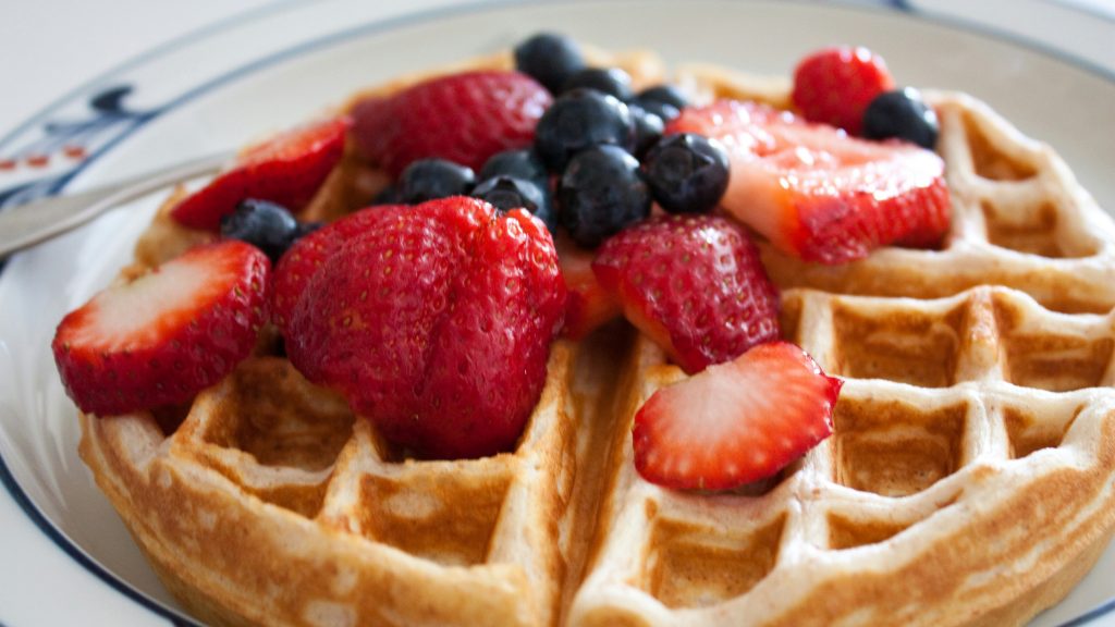 A close-up of a Country Waffles breakfast shows a golden-brown waffle topped with fresh strawberries and blueberries. The vibrant berries add a colorful contrast, and a fork rests beside the plate. Ideal for any Breakfast Club gathering.