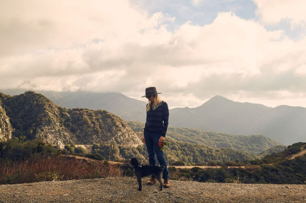 A person in a hat and striped sweater stands on a rocky path with their black dog. Mountains rise in the background under a cloudy sky, offering a clear image of Big Bear during fall. The scene features an outdoor, mountainous landscape and distant rolling hills, ideal for an active visit with your pet.