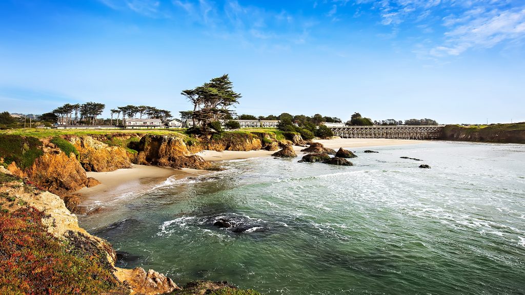 The North Coast shoreline offers a stunning view with its rugged cliffs, sandy beaches, and rocky outcrops. Near Fort Bragg, several houses are tucked among the trees in the background. A bridge spans across the water under a mostly clear sky with sparse clouds on the horizon.