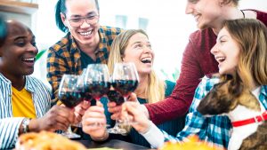 Five people sit around a table in a cozy Sacramento home, smiling and laughing as they raise glasses of red wine. A dog is also present, contributing to the lively scene.