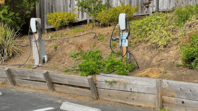 Two electric vehicle charging stations with coiled cords are mounted on wooden posts set in a sloped, grassy area beside a wooden fence. The parking space in front of the chargers is empty, featuring pavement interspersed with concrete barriers.