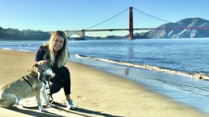 A person kneels on a sandy beach next to a dog with the Golden Gate Bridge visible in the background. It's a clear, sunny day, and gentle waves wash onto the shore beside them. Both look directly at the camera, capturing a moment by the water in San Francisco Bay Area.