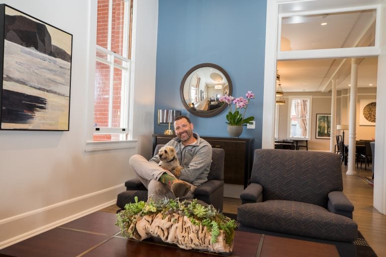 A person sits comfortably in a dimly lit chair within the warm, inviting atmosphere of the Lodge at the Presidio's living room. They hold a small dog affectionately, their smile reflecting contentment. The room is thoughtfully decorated, featuring a blue accent wall that adds depth and character. A round mirror hangs stylishly above a piece of art, adding to the aesthetic appeal. Sunlight filters through large windows, creating a natural ambiance. On the wooden table before them is an elegant arrangement of succulents, lending a fresh touch to the cozy setting.