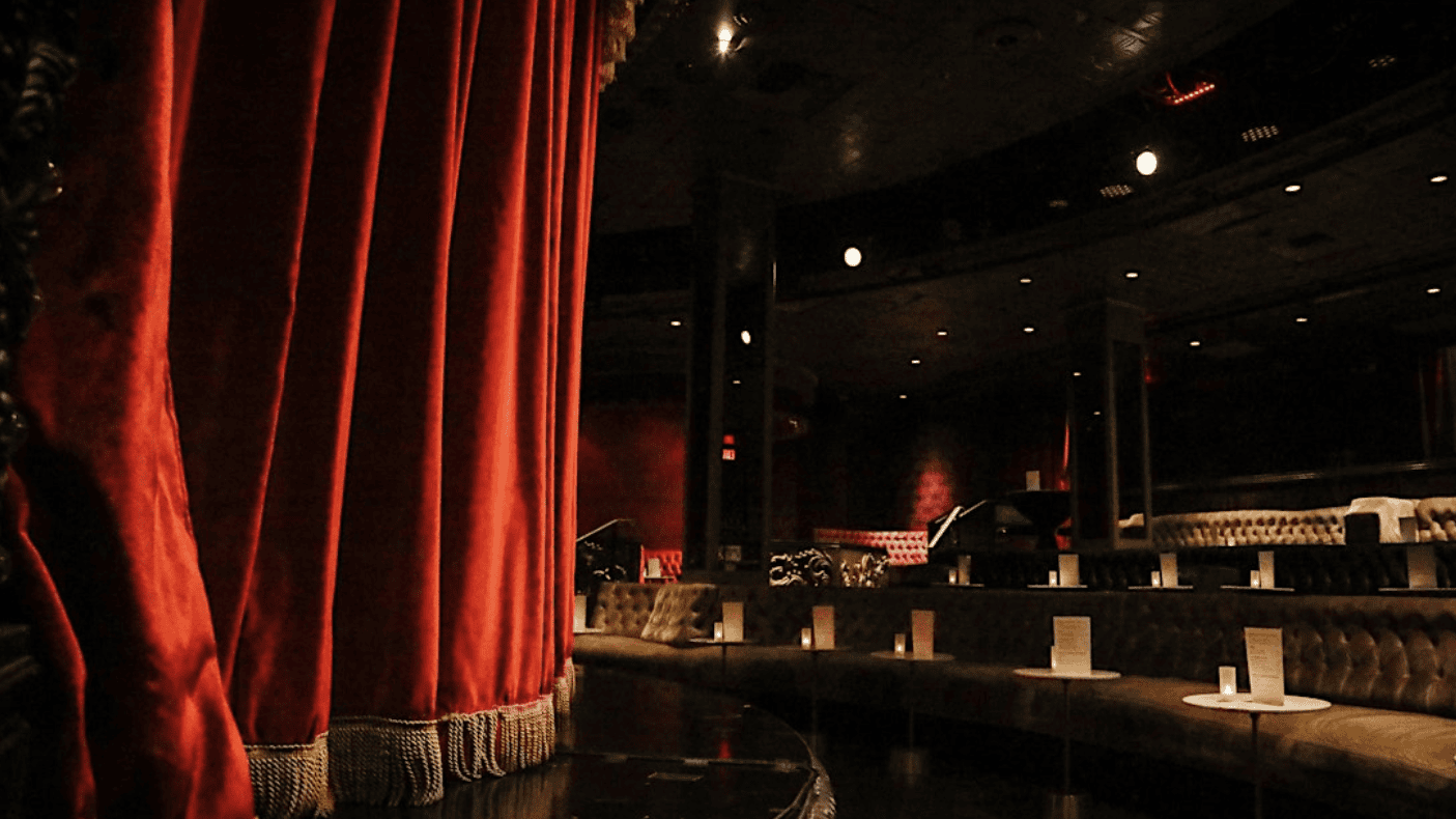 An empty theater with dim lighting and plush seats arranged in a semi-circle around a dark stage sets the scene for an evening of Can't Even Comedy. The vibrant red stage curtains stand out against the dark interior, while small tables with menus are placed in front of each seat.