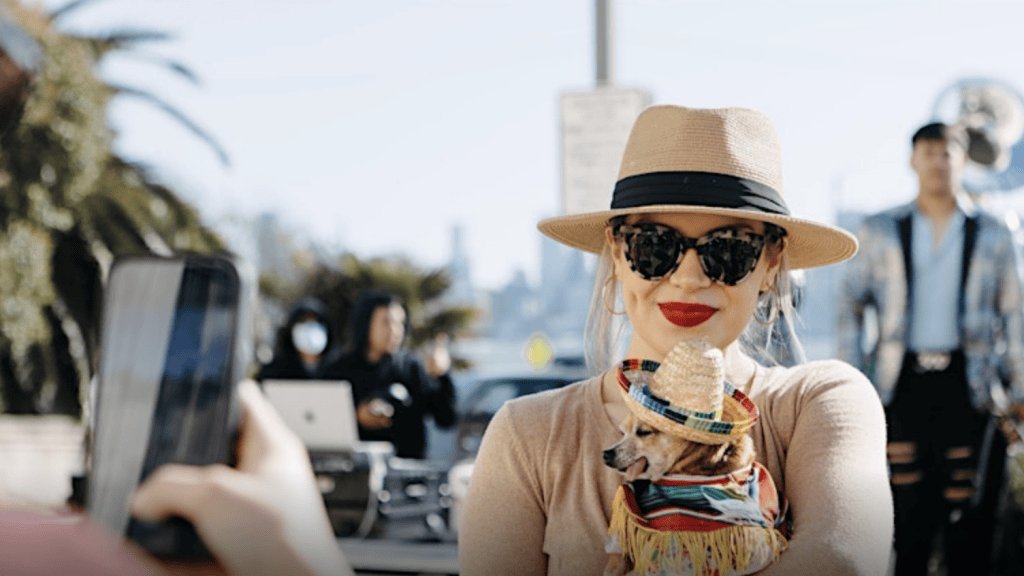 A woman in a straw hat and sunglasses holds a small dog dressed in a vibrant outfit and tiny sombrero. They are at a lively dog show. She smiles as another person takes their photo. Palm trees and people fill the background under clear, sunny skies.