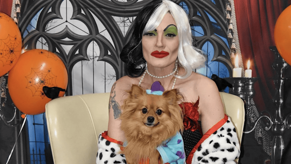 A person in a Dalmatian-patterned outfit and green eyeshadow holds a small brown dog dressed in a colorful costume. They are seated in front of a Gothic-style backdrop adorned with orange balloons and spiderwebs at the Wag Hotels' Haunted Howl-o-ween Party.