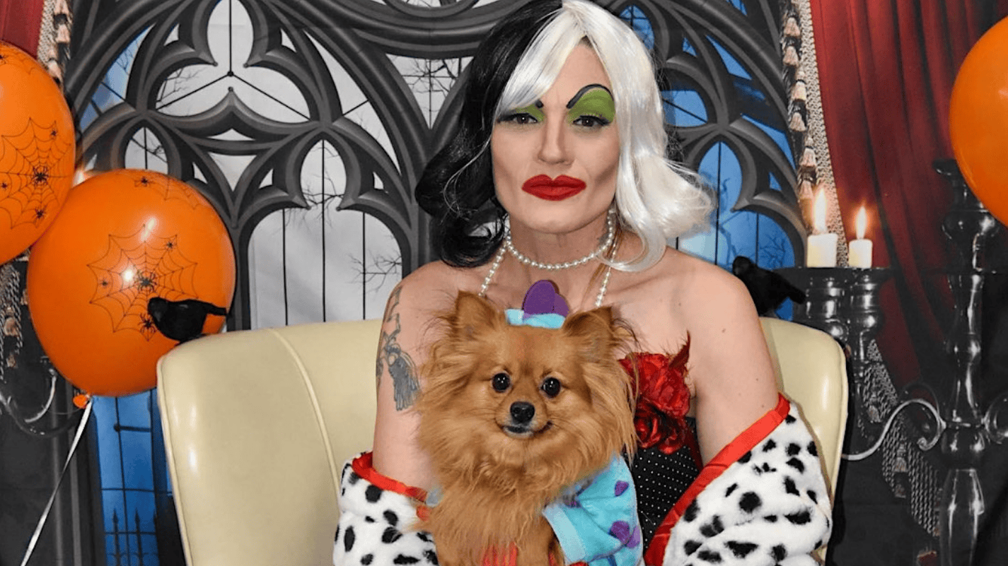 A person in a Dalmatian-patterned outfit and green eyeshadow holds a small brown dog dressed in a colorful costume. They are seated in front of a Gothic-style backdrop adorned with orange balloons and spiderwebs at the Wag Hotels' Haunted Howl-o-ween Party.