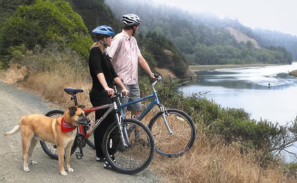 Two eco-minded individuals wearing helmets stand with their bikes on a dirt path next to a scenic river. Beside them is their tan dog, sporting a red bandana. The scene includes lush greenery and mist-covered hills in the background, creating a serene moment in nature.