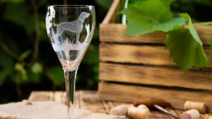 A wine glass etched with a dog design sits on a rustic wooden table. Around it are wine corks, a burlap cloth, and a wooden crate with grapevines. The blurred greenery in the background suggests the vineyards of California.