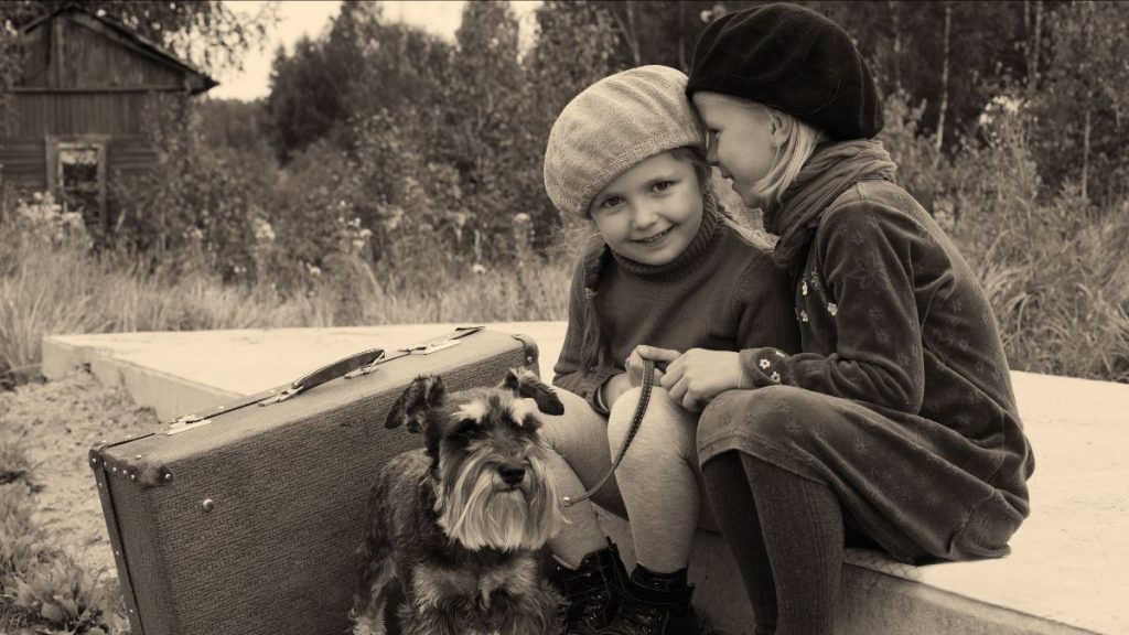 Two children in vintage outfits and berets sit on a bench beside an old suitcase in Mendocino County. One child whispers to the other as a small Schnauzer sits at their feet. Coastal greenery and an aged wooden building form the background of this sepia-toned photo.