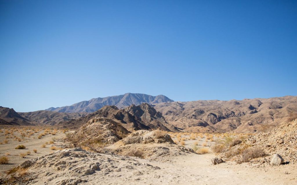 In Greater Palm Springs, a breathtaking desert landscape awaits exploration with your four-legged friend. The terrain is rocky, accented by sparse vegetation and gentle hills that rise subtly in the distance. Above, an endless blue sky stretches in all directions, providing a perfect contrast to the earthy hues below. A striking mountain range looms on the horizon, creating a stunning backdrop that invites adventure and discovery for both you and your dog as you wander through this expansive natural setting.