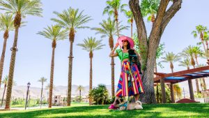 A woman stands on a grassy lawn beneath tall palm trees in Greater Palm Springs. She wears a colorful skirt, green top, and pink hat with sunglasses. She holds a black dog while two more dogs sit at her feet. The area has distant mountains and a clear blue sky.