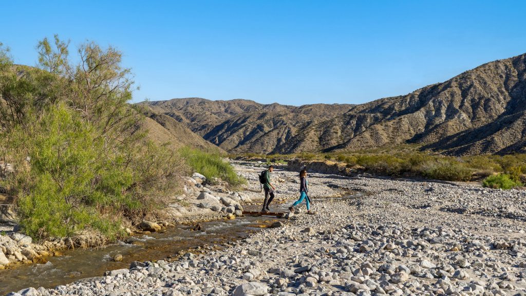 Two hikers move through a rocky, dry landscape with a shallow stream winding through it. Trails crisscross the terrain, which is flanked by rugged brown hills under a clear blue sky. Sparse plants hint at the desert setting, making it ideal for hiking with dogs.