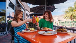 Two women sit at an outdoor restaurant table, toasting with cocktails and smiling. The table is set with waffles, pastries, and fruit. The sun shines brightly while palm trees and umbrellas create ample shade for a comfortable brunch experience.