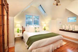 Discover the cozy attic bedroom at the Grape Leaf Inn, a pet-friendly retreat perfect for travelers with dogs. This inviting space features a comfortable double bed adorned with crisp white linens and green accents that create a fresh, harmonious feel. Stained glass windows add character and charm, while green lamps cast a gentle glow across the room. A cushioned bench offers a delightful spot to relax or curl up with your furry companion. The room's hardwood floors are practical for pets and give an elegant touch, complemented by soft lighting that enhances its welcoming ambiance.
