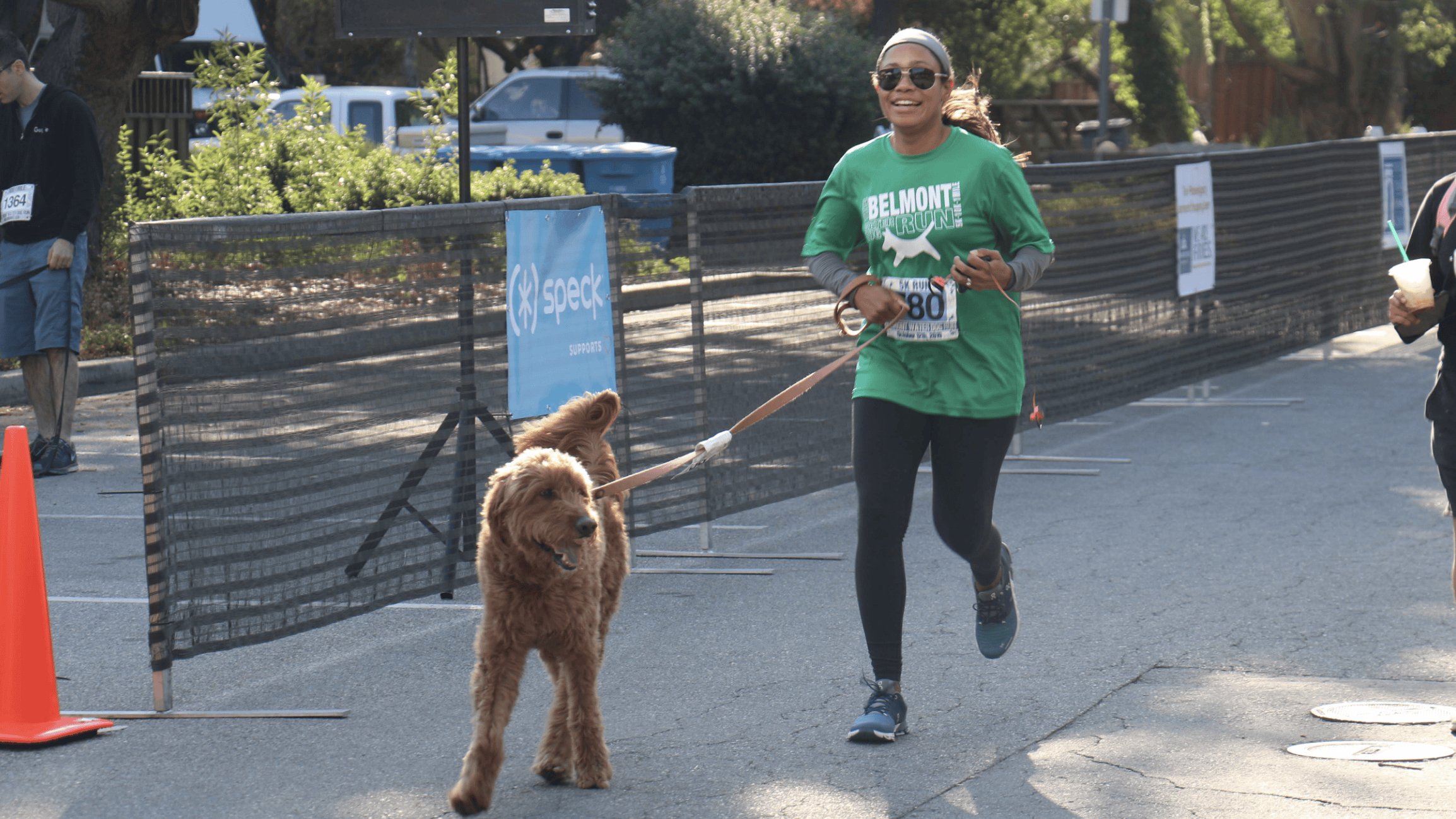 A woman in a green "Belmont" shirt and black leggings guides her fluffy brown dog on a leash as they participate in the Water Dog Run. The pair moves briskly along the road, sunglasses shielding her eyes from the sun. Behind them, an event sign stands near a sturdy barrier, marking their place along this popular race track for canine companions.