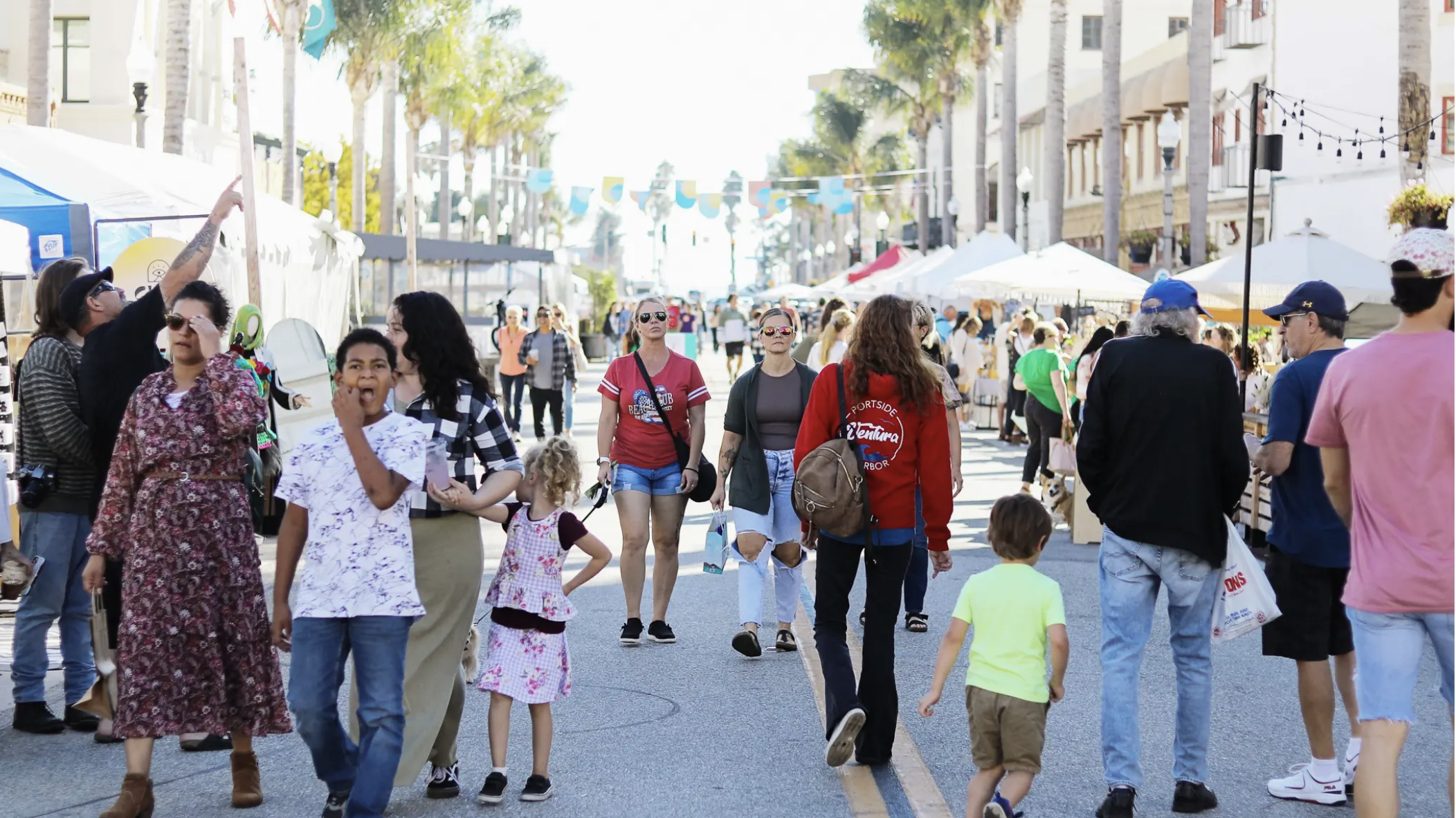 The street is lively and filled with the energy of people exploring a bustling fair. Tents stretch along the road, shaded by towering palm trees, each offering unique goods or tasty snacks. Among the crowd, you’ll find individuals dressed in relaxed attire and families navigating with children by their side. It’s a sunny day, perfect for strolling through this engaging community event with your dog.