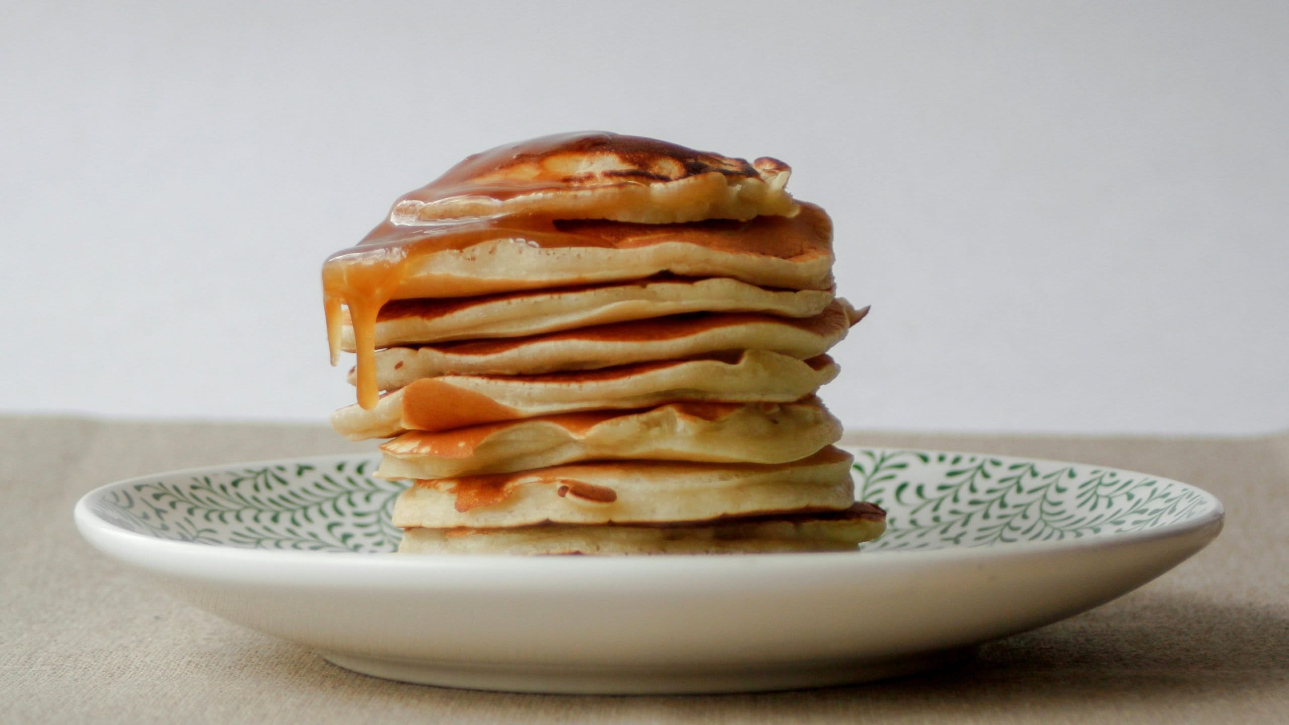 A stack of golden-brown pancakes, glistening with syrup as it drips down the sides, rests invitingly on a white plate adorned with a delicate green leaf pattern. This serene setting against a neutral beige background creates an inviting ambiance, perfect for enjoying Shelter Brunch while supporting the Butte Humane Society's fundraising event.