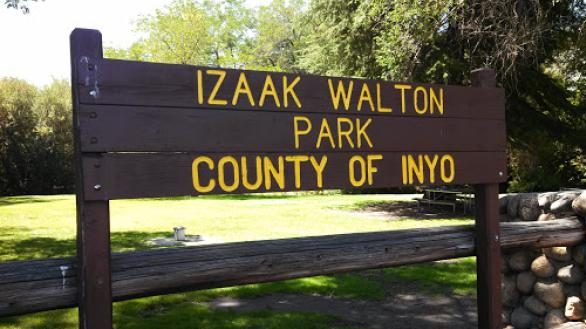 A brown wooden sign at the entrance reads "Izaak Walton Park County of Inyo." The sign stands on two posts beside a fence made of stone and wood. Behind it, green grass and mature trees indicate an open, shaded area suitable for walks or outdoor activities with dogs.
