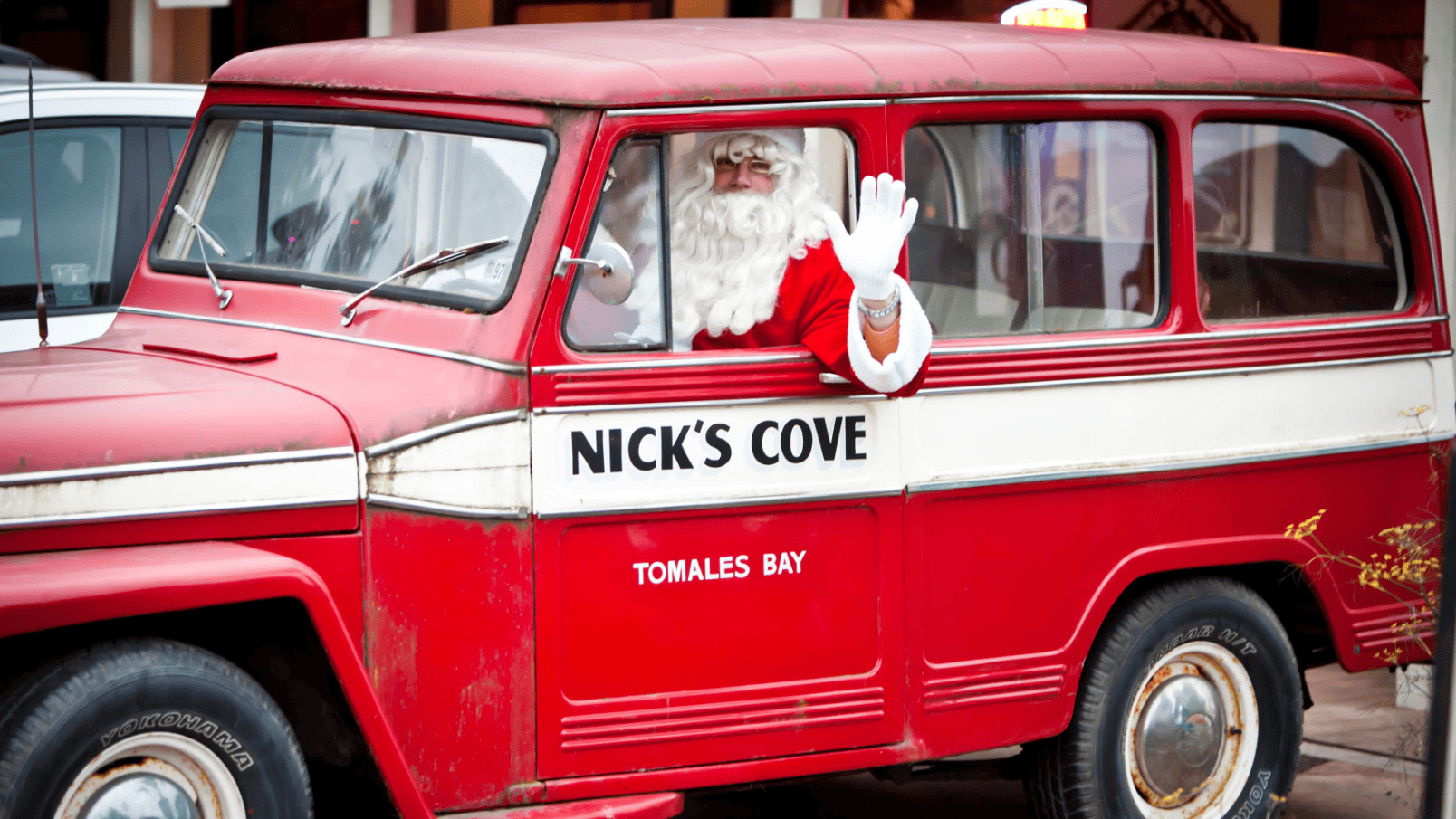 Santa Claus sits in the driver's seat of an eye-catching vintage red vehicle adorned with the words "Nick's Cove" and "Tomales Bay" on its side. The festive scene reflects the spirit of holiday cheer, making it an ideal setting for Santa Day celebrations.