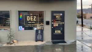 The Dez Fine Food shop features a welcoming street view characterized by its inviting window display. Through the glass, you can see handwritten menu items and an illuminated “Open” sign, signaling hospitality. Next to the window is a door marked with bold branding and the address 61705, clearly visible to passersby. A sandwich board stands nearby on the sidewalk, detailing enticing options such as coffee, smoothies, and ice cream — perfect for refreshment during your outing with your dog. In front of this scene are charming desert plants which add a touch of local flavor and warmth to the entrance.