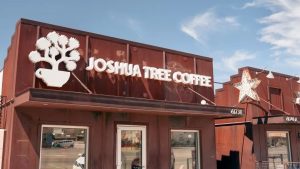 The coffee shop, with its rust-colored exterior, stands out with a sign that reads "Joshua Tree Coffee." The logo shows a coffee cup intertwined with a tree, hinting at the blend of nature and community offered inside. Beneath the wide-open blue sky, a star-shaped light fixture catches the eye, adding a unique touch to this welcoming spot. It's an appealing place to relax and enjoy time with your dog by your side.