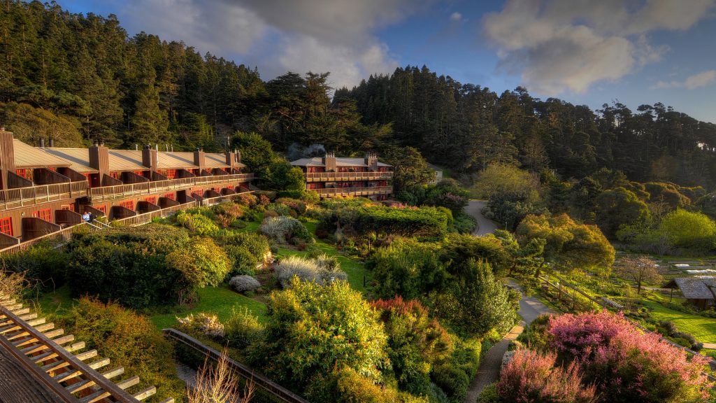 A Northern California hotel sits quietly among hills blanketed with dense, green forests. Its architecture includes balconies that overlook colorful gardens and thick clusters of trees, all under a sky dotted with clouds. This hotel is highly recommended by Condé Nast Traveler for its tranquil setting, where winding pathways meander through the lush landscape.