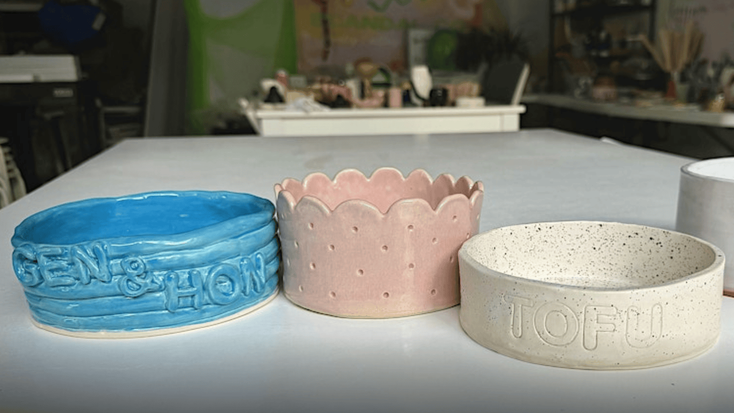 Three handmade ceramic bowls sit on the table. On the left, a blue bowl bears the names "GEN & HON." In the middle, there is a pink bowl with scalloped edges. The last bowl on the right is white with speckles and has "TOFU" embossed on its surface. These creations are part of a dog-friendly pottery workshop at Woofs & Clay, where participants and their four-legged friends explore their artistic sides together.