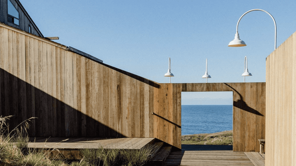 At Sea Ranch Lodge, a coastal wooden deck provides a stunning setting for dog walkers and their canine companions. The deck features four suspended white pendant lights, their soft illumination creating intricate patterns on the walls as evening approaches. Framing an uninterrupted view of the vast ocean under a clear blue sky, the platform offers a peaceful space where you and your dog can enjoy the calming rhythm of rolling waves. In the foreground, patches of grass add a touch of green and invite dogs to explore or rest in between bouts of playfulness.