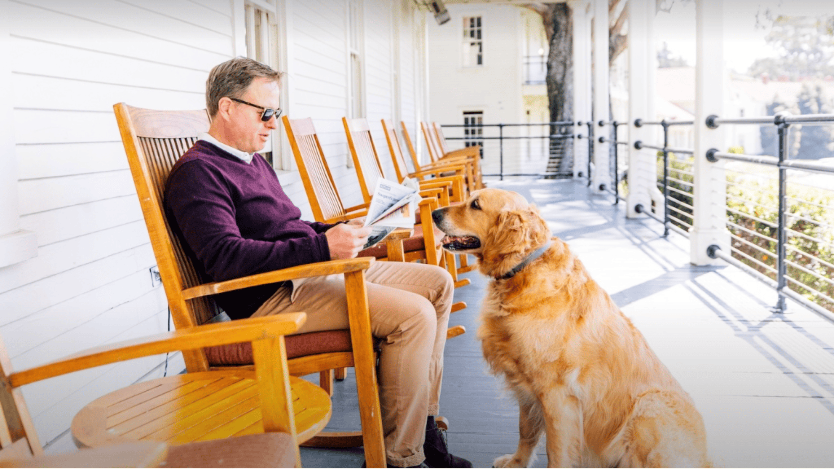 A man wearing sunglasses sits on the porch at Cavallo Point Lodge, engrossed in a newspaper while gently swaying in a wooden rocking chair. In front of him, a golden retriever sits attentively, gazing up at him. The porch is characterized by its white wooden walls and an array of rocking chairs arranged neatly for guests to use.