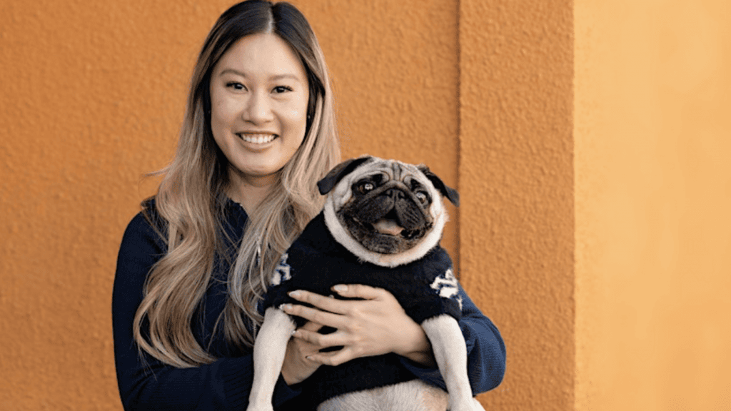 A smiling individual with long hair cradles a content pug wearing a dark sweater. Both are poised for Pupsgiving festivities, standing against an orange textured wall.