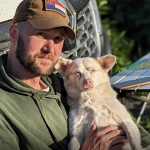 A man wearing a green hoodie and a brown cap sits outside with his small white dog, a moment of quiet amidst the bustling festival activities. Next to them, a vehicle and foldable table provide a makeshift resting spot. The area is lush and green, reflecting the lively environment of Paws For Ukraine.