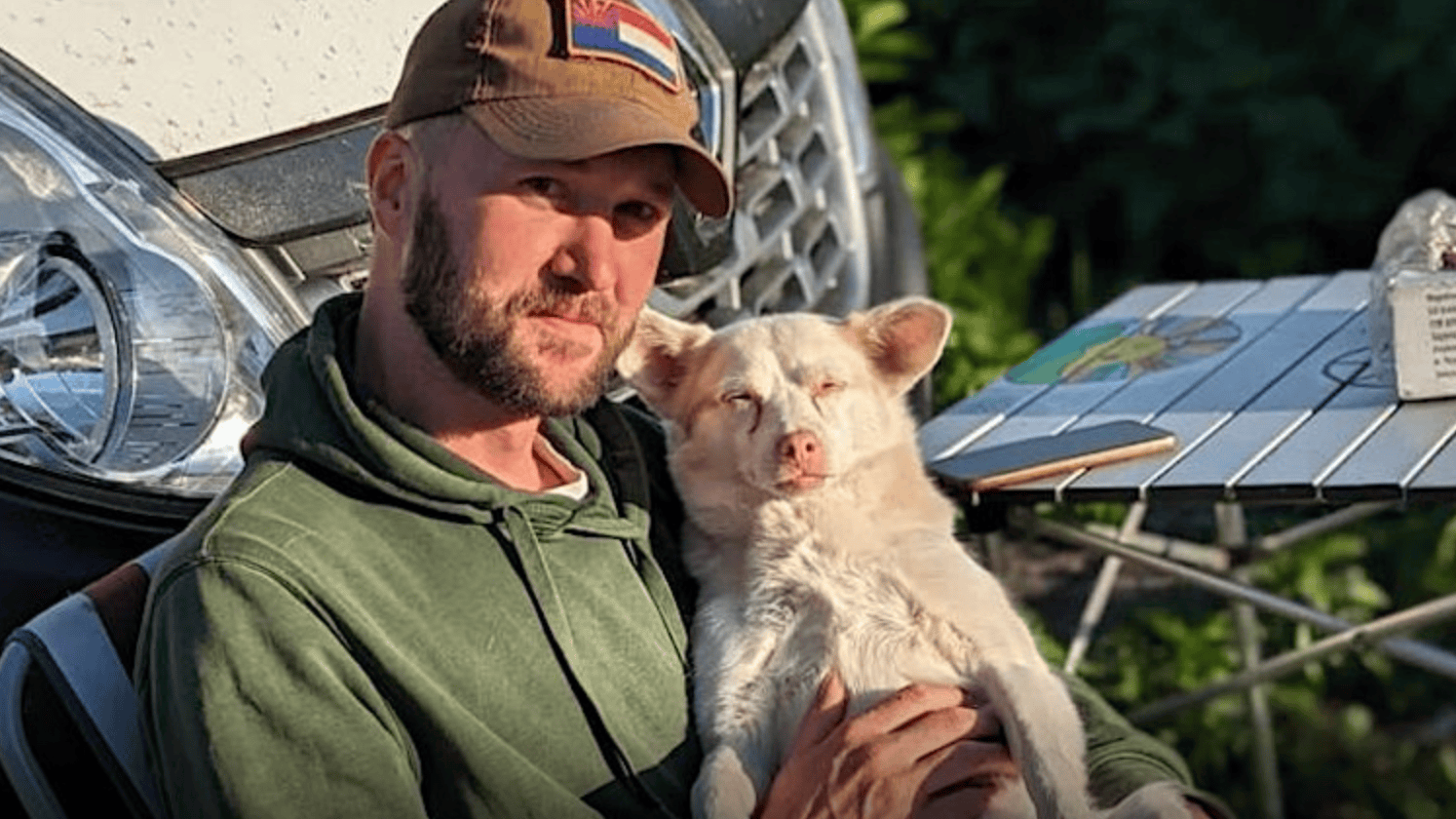 A man wearing a green hoodie and a brown cap sits outside with his small white dog, a moment of quiet amidst the bustling festival activities. Next to them, a vehicle and foldable table provide a makeshift resting spot. The area is lush and green, reflecting the lively environment of Paws For Ukraine.