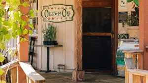 A quaint storefront with a wooden deck displays a sign for "Temecula Olive Oil Company." Painted olive branches decorate the entrance, complemented by lush potted plants. Near a rustic wooden barrel, an "Open" sign warmly welcomes visitors to explore a carefully curated selection of premium olive oils.