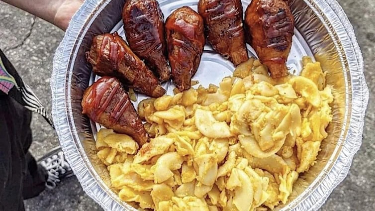 A foil tray holds a collection of BBQ baked chicken drumsticks, each piece grilled to a deep, inviting glaze. Alongside sits a portion of creamy macaroni and cheese, its golden hue promising rich flavor. This combination makes for an ideal dish at any casual outdoor event with your dog by your side.