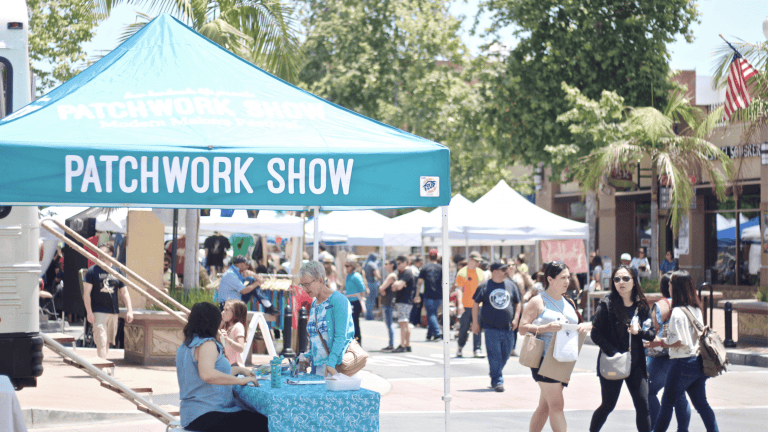 A vibrant dog park bathed in sunlight features abundant greenery with mature trees, offering ample shade for leisurely outdoor activities. In the distance is a well-equipped playground that presents an additional activity area. An inviting picnic table spreads out nearby, ideal for enjoying meals al fresco or taking relaxing breaks from play. A neatly-placed trash bin aids in maintaining the park's cleanliness for everyone's enjoyment. Dominating the scene is a generously wide dirt path gracefully curving through the expanse of grass, promising exploratory walks and lively fetch games.