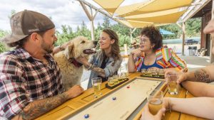 In the outdoor seating area of a cozy dining spot near Mariposa County, four people are gathered around a table, their conversation mingling with the cheerful ambiance. A large dog lounges contentedly at their feet, enjoying gentle pats from one of the individuals. Overhead, a canopy provides welcome shade from the sun's rays. On the table and nearby surfaces rest several glasses of beer, catching glints of sunlight in their amber liquid. Off to one side stands a shuffleboard table, inviting both humans and canines to enjoy this relaxed setting where good company is effortlessly shared alongside man's best friend.